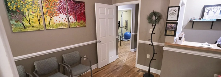 Waiting room with three chairs under colorful tree paintings, an open door showing exercise equipment, and a reception desk with certificates and decorations.