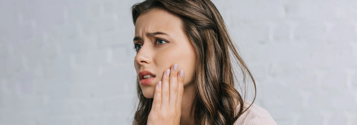 Person with long wavy brown hair resting a hand on the chin, wearing a light-colored top, with a neutral brick wall background.