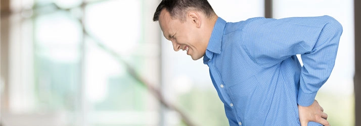 Person wearing a blue checkered shirt leaning forward and holding lower back, illustrating discomfort or pain.