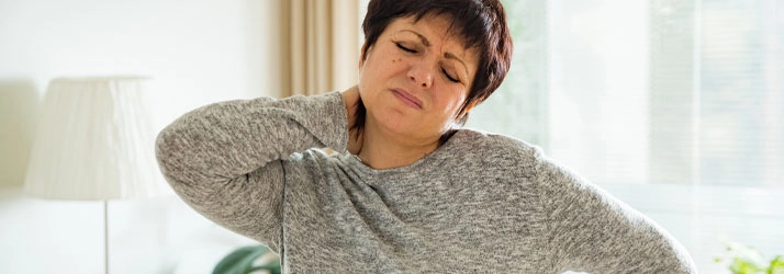 A person wearing a long-sleeved gray sweater is indoors, standing with one hand placed on the back of the neck and the other on the hip, suggesting discomfort or strain.