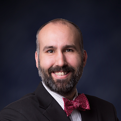 Professional headshot of a bald man with a full beard, dressed in a dark suit, white shirt, and red patterned bow tie, against a dark blue background.
