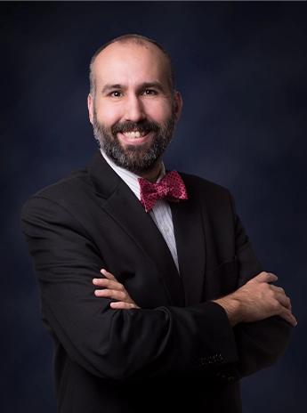 Person in a dark suit with a white shirt and red bow tie, smiling with arms crossed against a plain dark background.